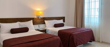 Habitación de hotel con dos camas, cabecera de madera, mesita de noche, lámpara y ventana con cortinas.