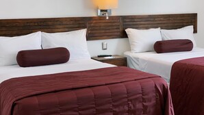 Habitación de hotel con dos camas, cada una con sábanas blancas y almohadones color burdeos. Tiene un cabecero de madera, una mesita de noche con lámpara y una lámpara de pared.