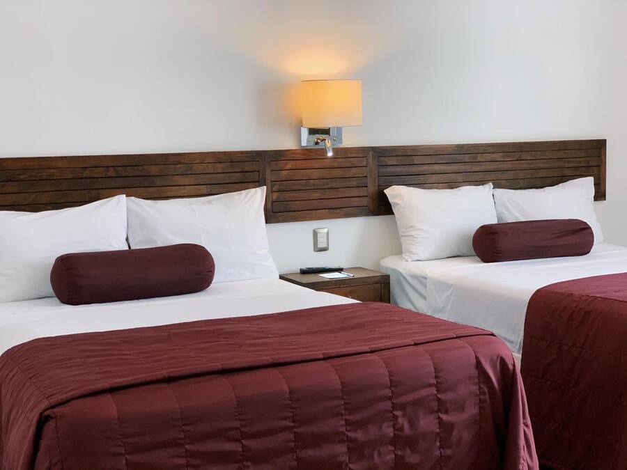 Habitación de hotel con dos camas, cada una con sábanas blancas y almohadones color burdeos. Tiene un cabecero de madera, una mesita de noche con lámpara y una lámpara de pared.