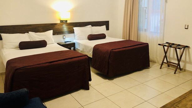 Una habitación de hotel con dos camas, una mesita de noche, una lámpara, una ventana con cortinas y una pequeña mesa.