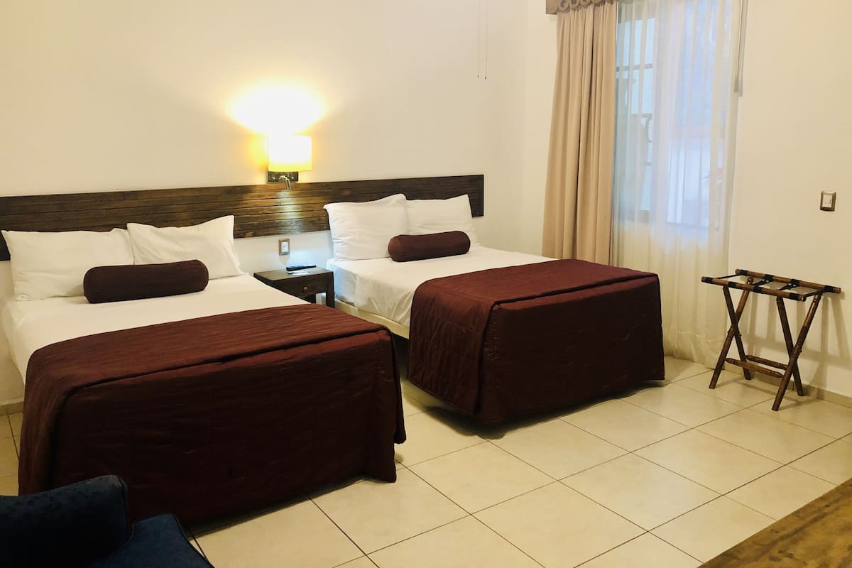 A hotel room with two beds, a nightstand, a lamp, a window with curtains, and a small table.