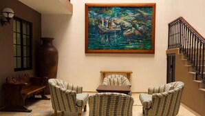 Una habitación con sillones estampados, un banco de madera, un jarrón grande y un cuadro de un paisaje fluvial.