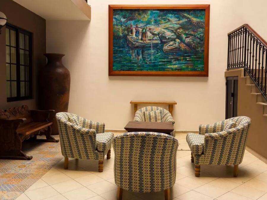 Una habitación con sillones estampados, un banco de madera, un jarrón grande y un cuadro de un paisaje fluvial.