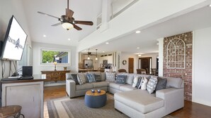 A spacious living room with a sectional sofa, a round coffee table, and a television. A kitchen is visible in the background with a dining area.