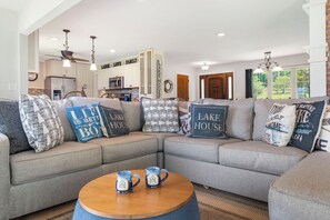 Una sala de estar moderna con un sofá seccional decorado con almohadas 'Lake House', una mesa de centro de madera con tazas, y una cocina al fondo.