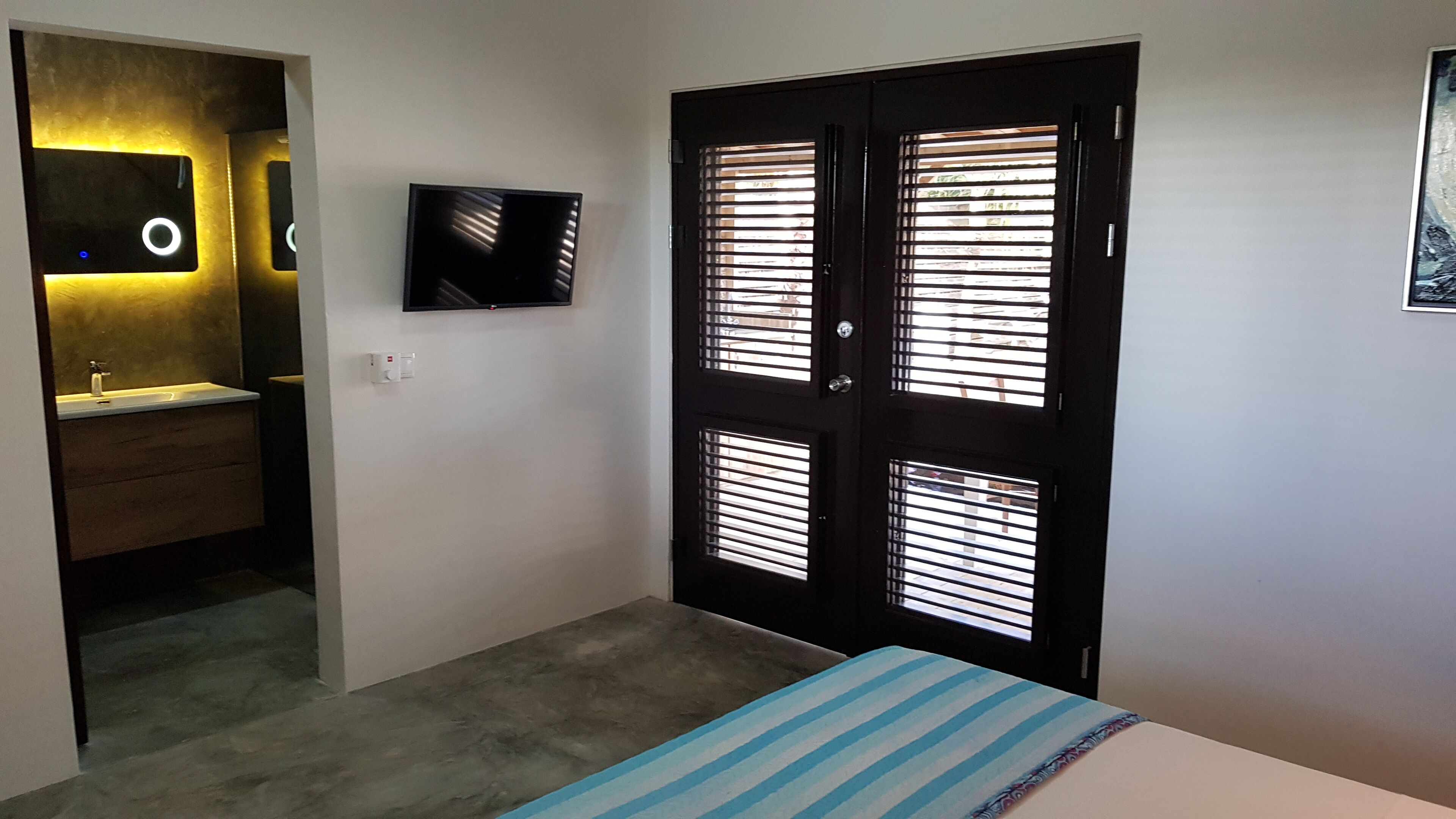 A bedroom with a bed, a TV, a door with blinds, and a bathroom with a sink and mirror.