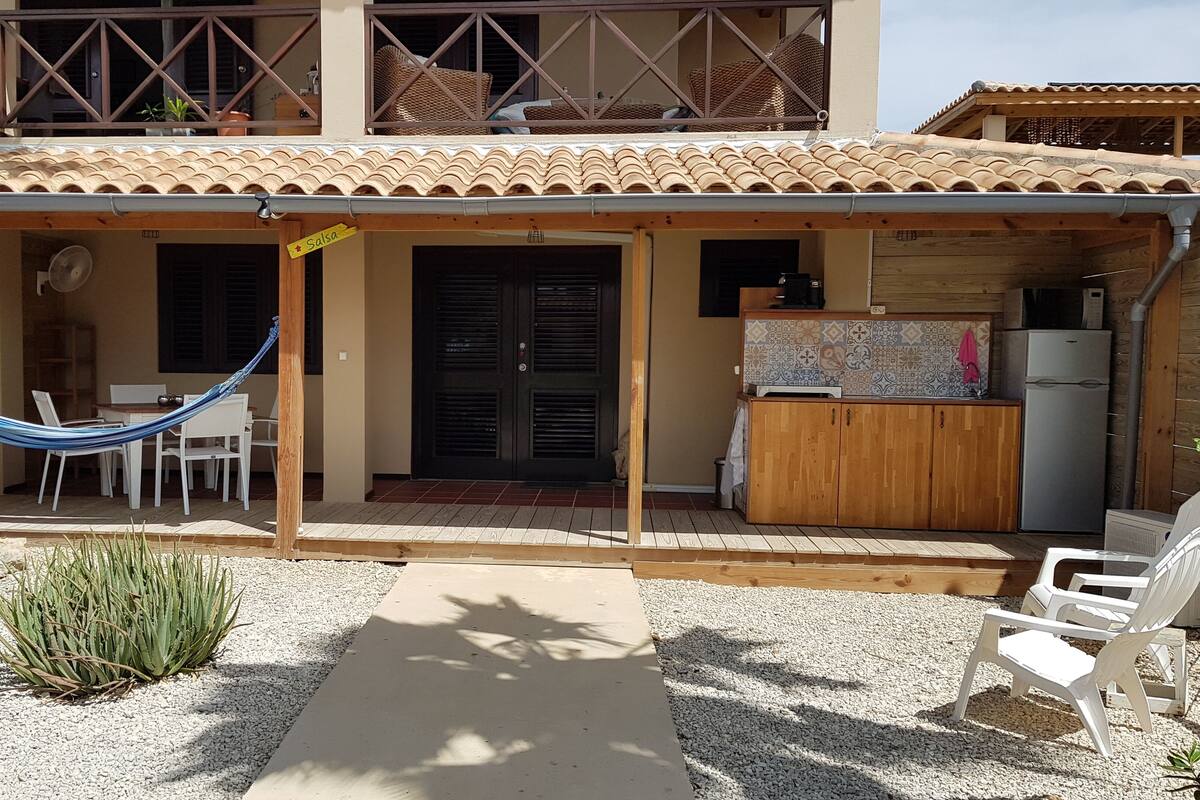 A two-story building with a tiled roof, a covered patio with white plastic chairs, a hammock, and a kitchen area with a refrigerator and a coffee maker.