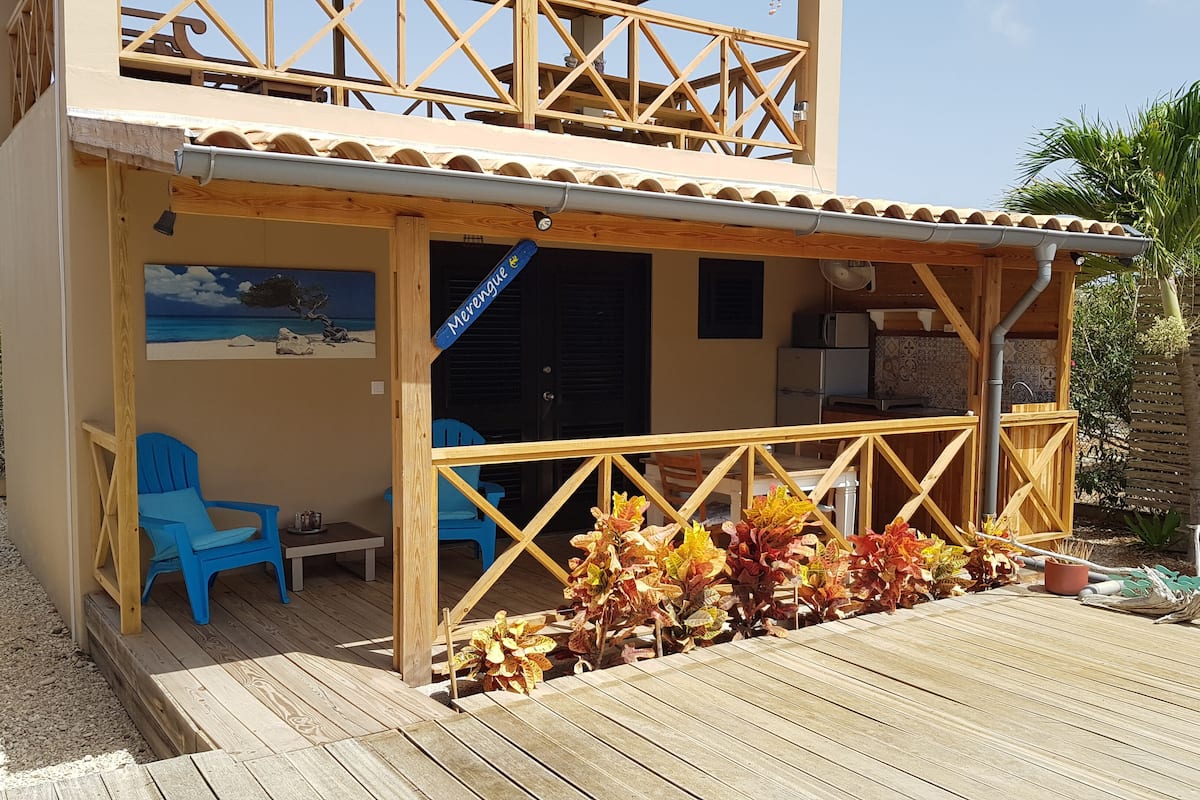 A two-story building with a wooden deck, a covered seating area with blue chairs, and a mural of a beach scene.
