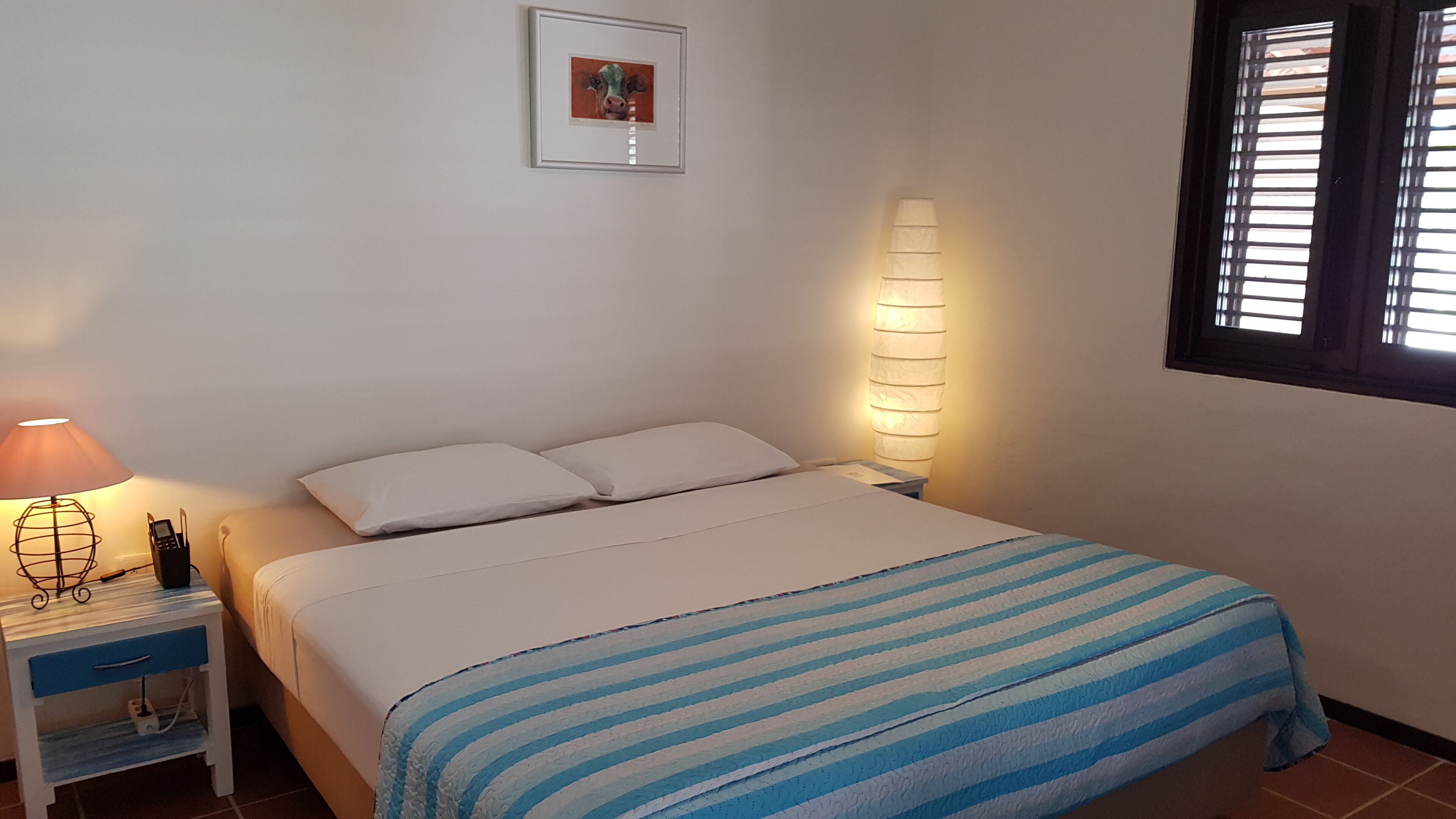 Un dormitorio con una cama, una mesita de noche con lámpara, un cuadro enmarcado en la pared y una ventana con persianas.