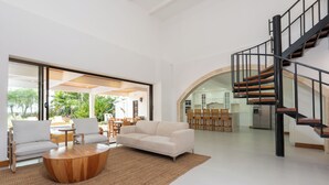 A spacious living area with a white sofa, a wooden coffee table, and a large window overlooking a garden. A spiral staircase is visible in the background.