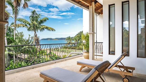 A balcony with two wooden lounge chairs, a view of the beach, and palm trees.