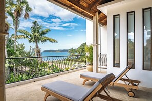 A balcony with two wooden lounge chairs, a view of the beach, and palm trees.