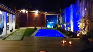 A nighttime outdoor pool area with a wooden deck, two lantern-style lights, and a blue-lit water feature. - Casa BluMarino (Porto Seguro)