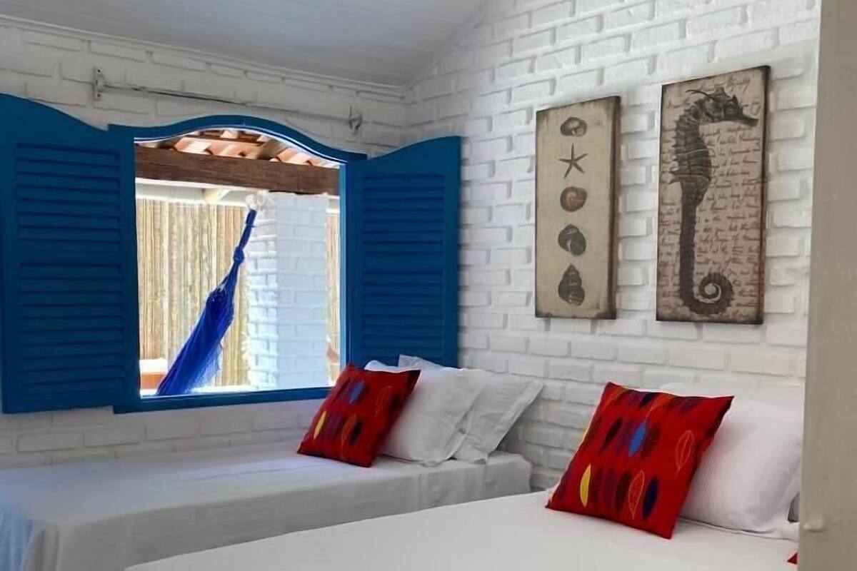 A room with two beds, white bedding, red pillows, a hammock, and a white brick wall with a seahorse artwork.