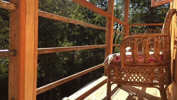 Una terraza de madera con una silla de mimbre, rodeada por una barandilla de madera, con vistas a un bosque frondoso.