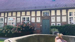 Een traditioneel vakwerkhuis met een opvallende groene deur, een stenen waterput en een tuin met roze bloemen.