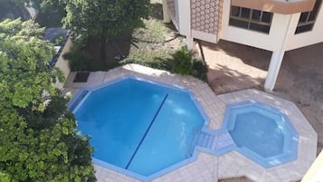 Un espace piscine sur le toit, comprenant deux piscines, entouré d’arbres et bordé d’un immeuble doté d’un balcon.