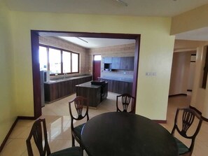Un coin repas avec une table ronde et quatre chaises, un espace cuisine avec des meubles sombres, et une vue sur un balcon à travers une porte ouverte.