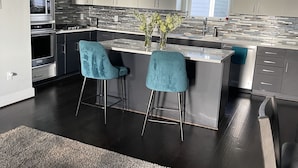 Une cuisine moderne dotée d’un îlot central, de deux tabourets de bar et d’une grande fenêtre qui laisse entrer la lumière naturelle.