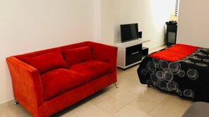 Une pièce avec un canapé rouge, un lit aux motifs noir et blanc, un téléviseur posé sur un meuble blanc et une petite armoire noire.