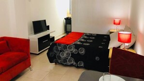 Une chambre d’hôtel de taille modeste, comprenant un lit, un téléviseur, un canapé rouge, une petite table avec un verre et une lampe rouge.