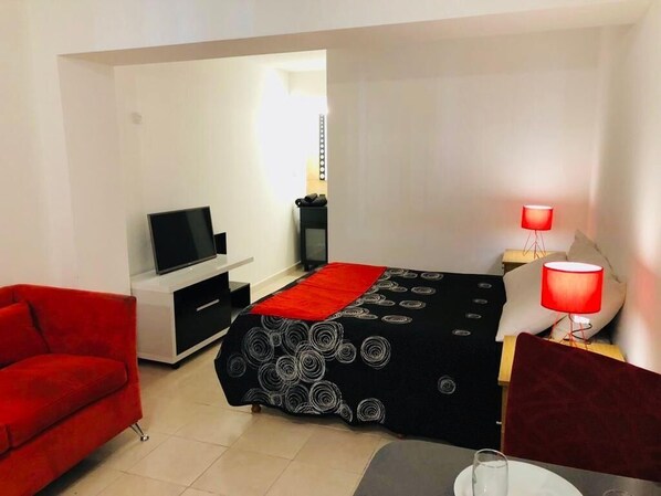 A compact hotel room with a bed, a TV, a red sofa, a small table with a glass, and a red lamp.