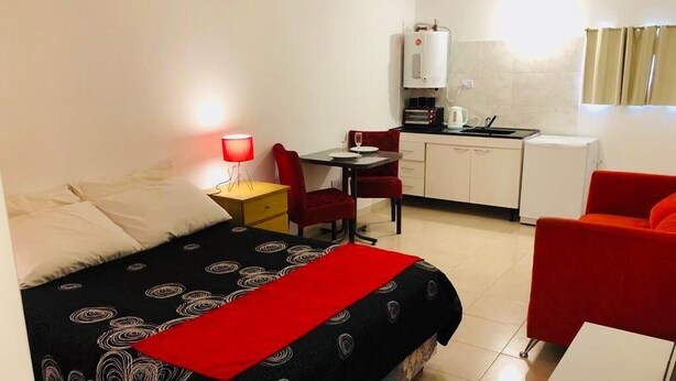 Une chambre d’hôtel compacte comprenant un lit, un bureau avec une chaise, une kitchenette et un fauteuil rouge.
