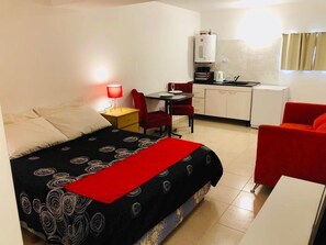 A compact hotel room with a bed, a desk with a chair, a kitchenette, and a red armchair.