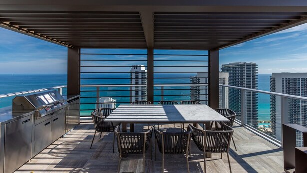 A balcony with a grill, dining table, and chairs, overlooking the ocean and high-rise buildings. - HYDE HOUSE NEW (Hollywood)