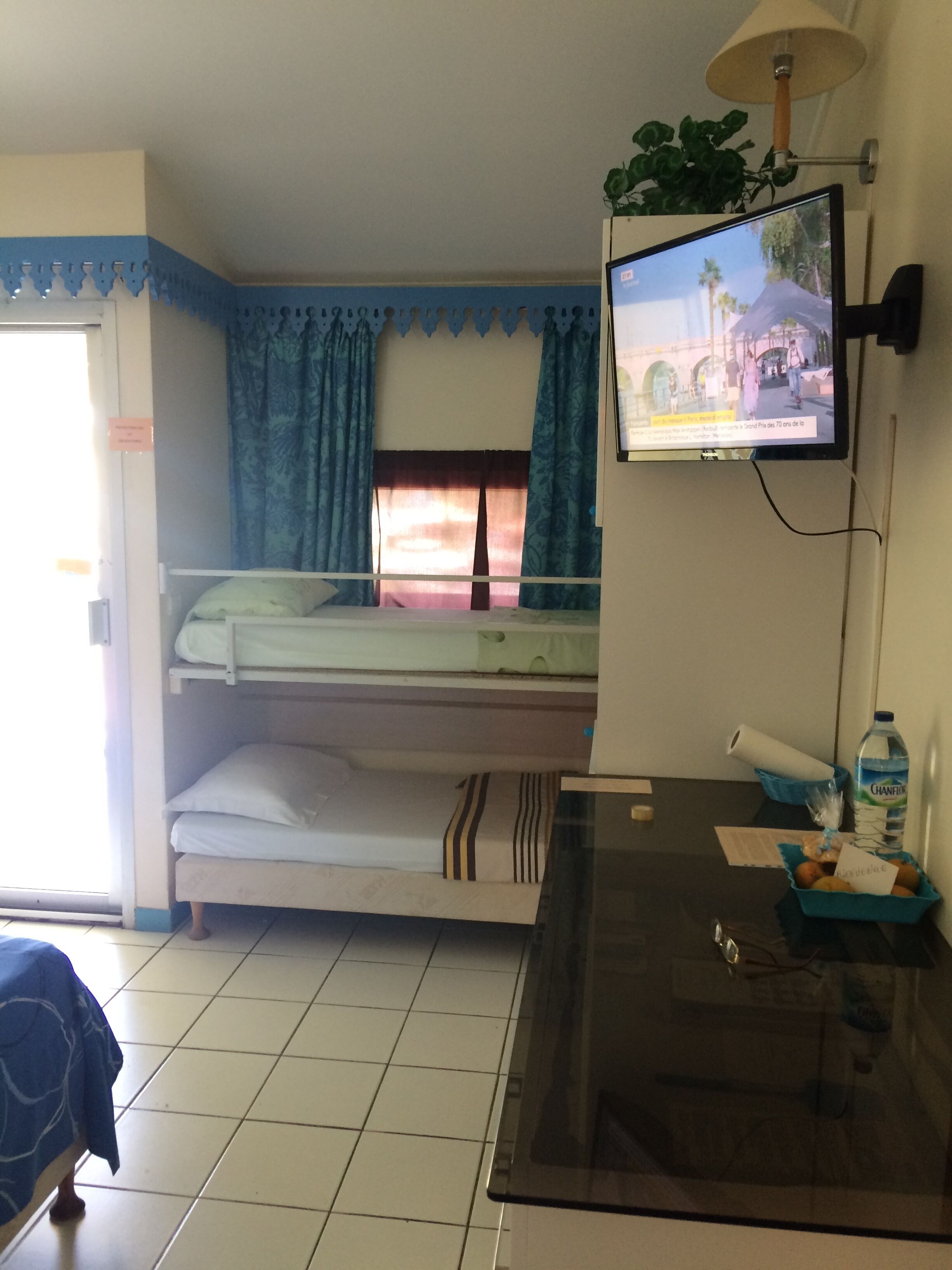 Ein Hotelzimmer mit Bett, Wandfernseher, kleinem Tisch mit Wasserflasche und einem Fenster mit Vorhängen.