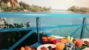 A table with breakfast items including fruits, a drink, and a cup of coffee, overlooking a body of water. - Beautiful, air-conditioned studio in a 3-star hotel: 2 pools + INTERNET, right on the water (Les Trois-Îlets)