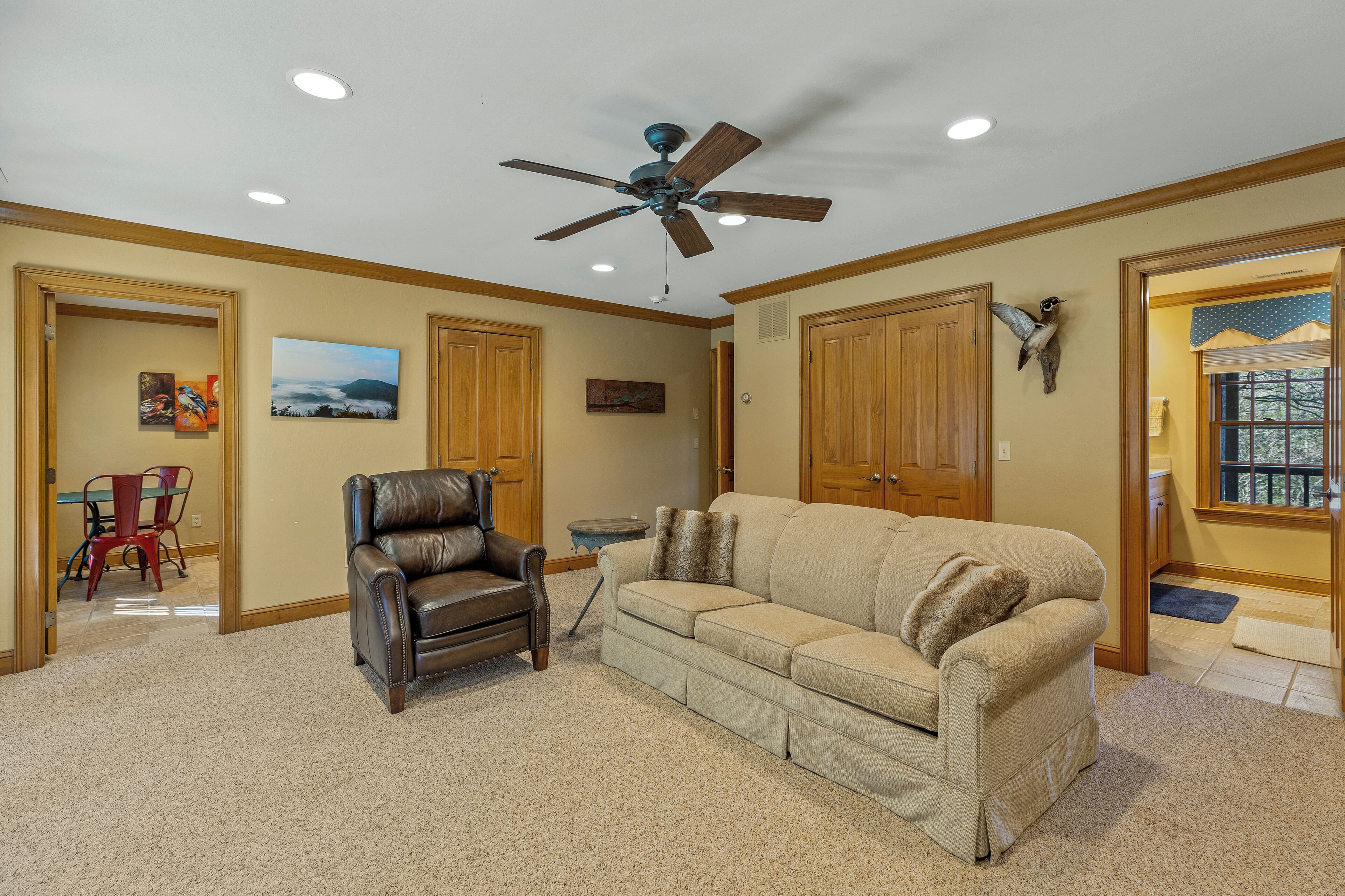 A living room with a ceiling fan, leather armchair, beige sofa, wooden doors, and a dining area with red chairs.