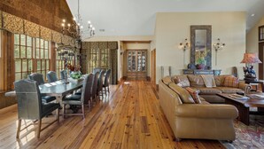 A spacious living area with a wooden floor, a dining table with chairs, a leather sofa, and a large mirror. - Fawn Creek: Beautifully Appointed, Mtn Views w Covered Porch and Wood Fireplaces (Highlands)