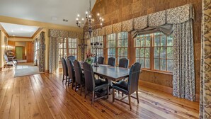 A dining room with a wooden table and chairs, large windows with patterned curtains, and a chandelier. - Fawn Creek: Beautifully Appointed, Mtn Views w Covered Porch and Wood Fireplaces (Highlands)