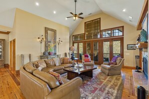 A spacious living room with a large sectional sofa, a fireplace, and a view of the outdoors through large windows. - Fawn Creek: Beautifully Appointed, Mtn Views w Covered Porch and Wood Fireplaces (Highlands)