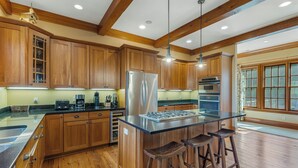 A spacious kitchen with wooden cabinets, a stainless steel refrigerator, a large island with stools, and a window providing natural light. - Fawn Creek: Beautifully Appointed, Mtn Views w Covered Porch and Wood Fireplaces (Highlands)