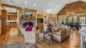 A spacious living area with a fireplace, wooden flooring, and a dining area with a large window. - Fawn Creek: Beautifully Appointed, Mtn Views w Covered Porch and Wood Fireplaces (Highlands)