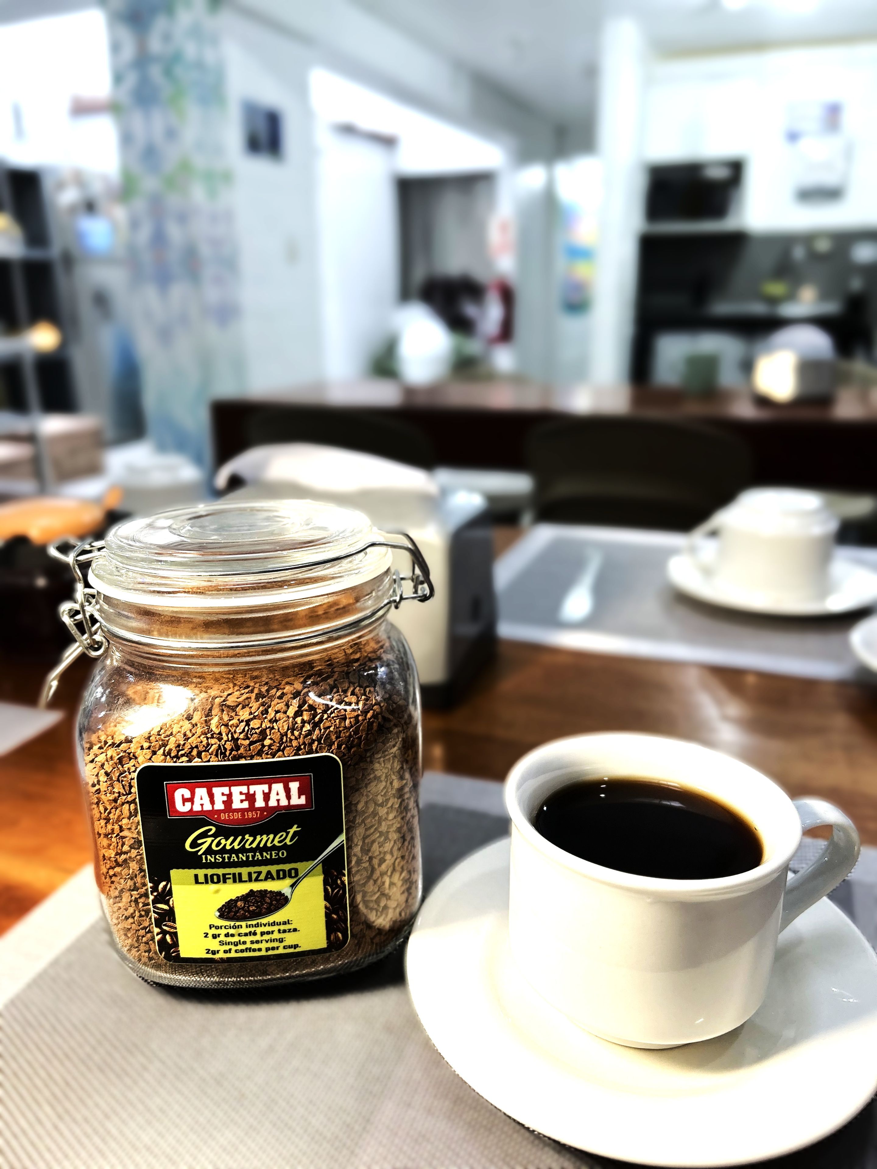 A jar of Cafetal Gourmet instant coffee and a cup of coffee on a table in a cafe setting.
