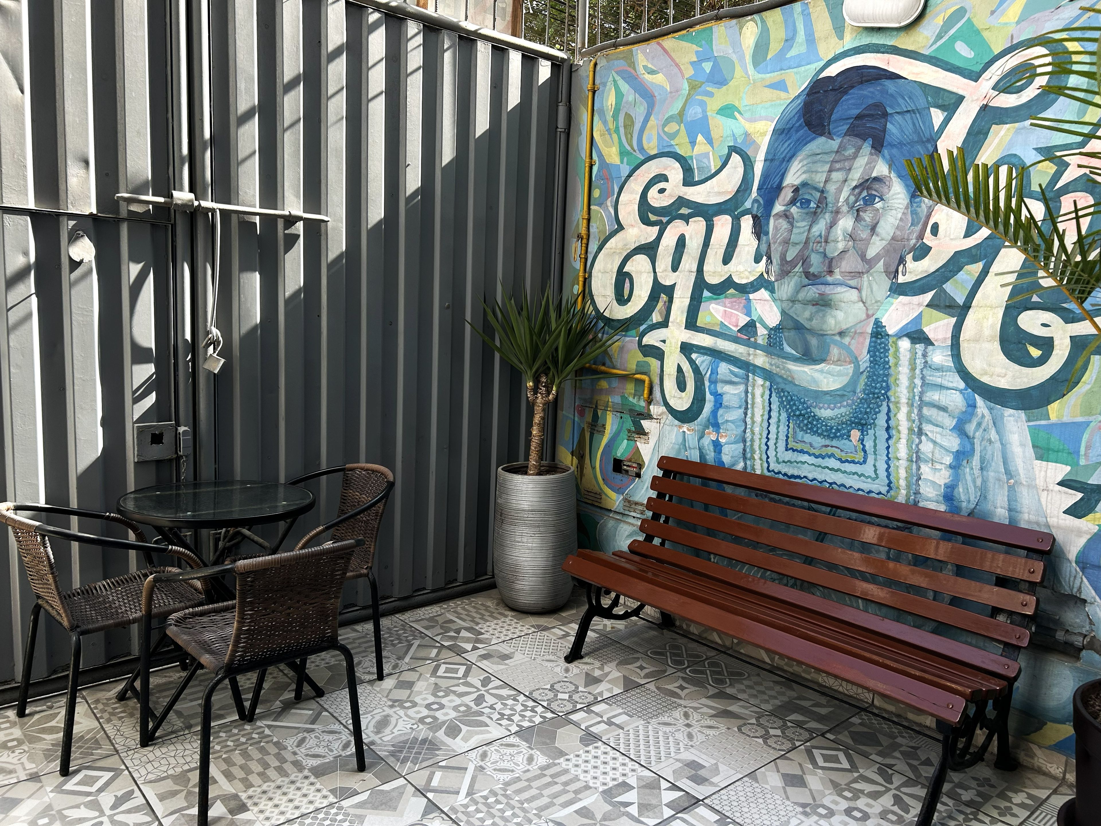 Un espace patio agrémenté d’une clôture en métal, d’une table avec des chaises, d’un banc et d’une fresque colorée sur le mur.