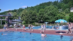 Une piscine entourée de personnes, un toboggan aquatique et une colline avec des arbres en arrière-plan.