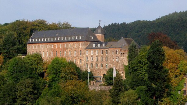 Un grand bâtiment en pierre, doté de nombreuses fenêtres et d’une tour, entouré d’arbres.
