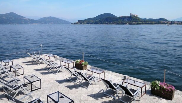 Een terras aan het water met ligbedden en plantenbakken, met uitzicht op een rustig meer en heuvels in de verte.