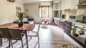 Een moderne keuken met een eethoek, met een houten tafel, stoelen, een klein rond tafeltje met bloemen, en uitzicht op groen via een raam.