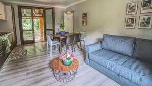 Een woonkamer met een grijze bank, een eettafel met stoelen, een keuken met kasten, en een groot raam met uitzicht op groen.