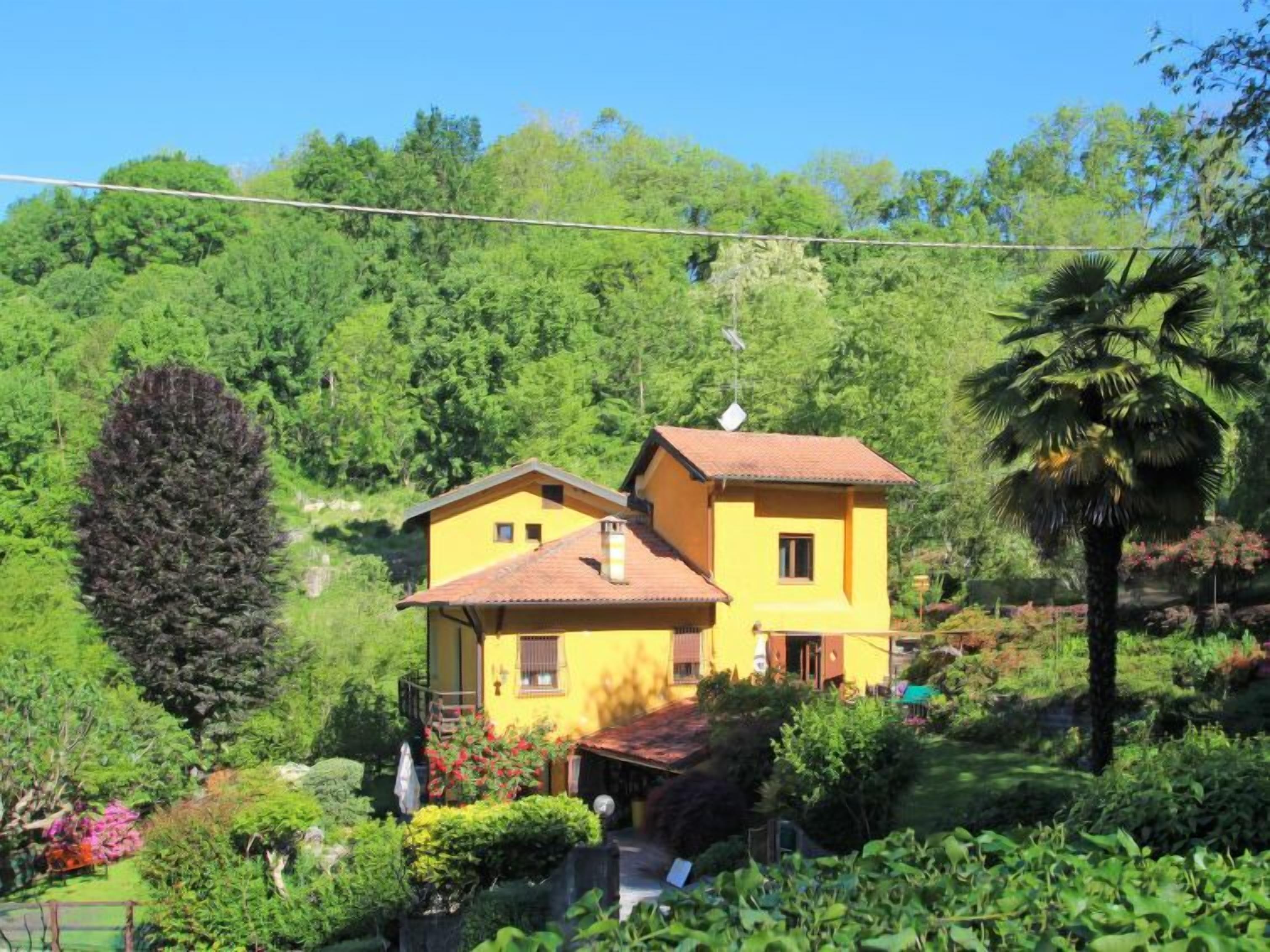 A two-story yellow house with a red roof, surrounded by lush greenery and a tall tree to the left.