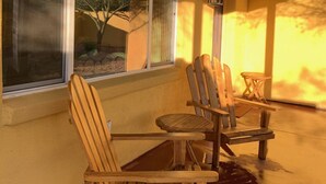 Un patio cubierto con sillas Adirondack de madera y una mesita, un ventanal con vistas a un árbol, y un aplique de pared.