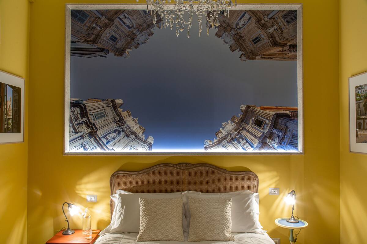 Quarto de hotel com uma grande imagem emoldurada de edifícios, uma cama com lençóis brancos, duas luminárias de cabeceira e um candelabro.