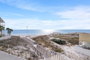 A beach view with a wooden boardwalk, a flag, and a building with a balcony overlooking the ocean.