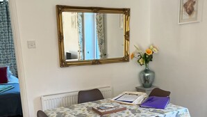 Un comedor con una mesa cubierta con mantel floral, rodeada de sillas marrones. Un jarrón con flores está sobre la mesa, y un espejo enmarcado cuelga de la pared de arriba.