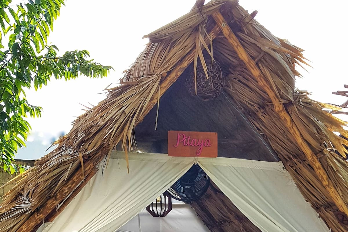 Una caba ña con techo de paja, un letrero que dice "Pitaya", una cama con un colorido edredón y una silla de madera.